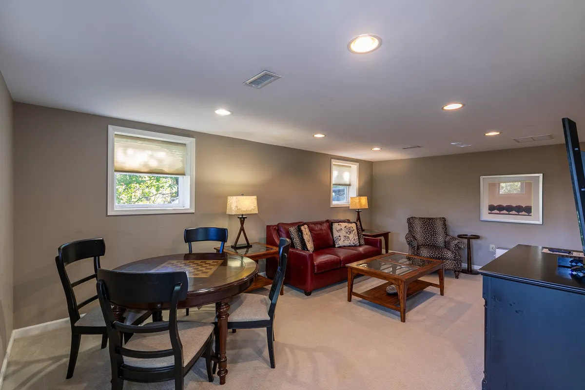 The image presents a welcoming interior with a red leather sofa, round dining table, TV, decorative lamp, and framed artwork. The arrangement suggests a multi-functional space where guests can enjoy casual gatherings, family game nights, or cozy evenings watching a movie. The mood is relaxed and comfortable, underlining the versatility of the entertainment zones.