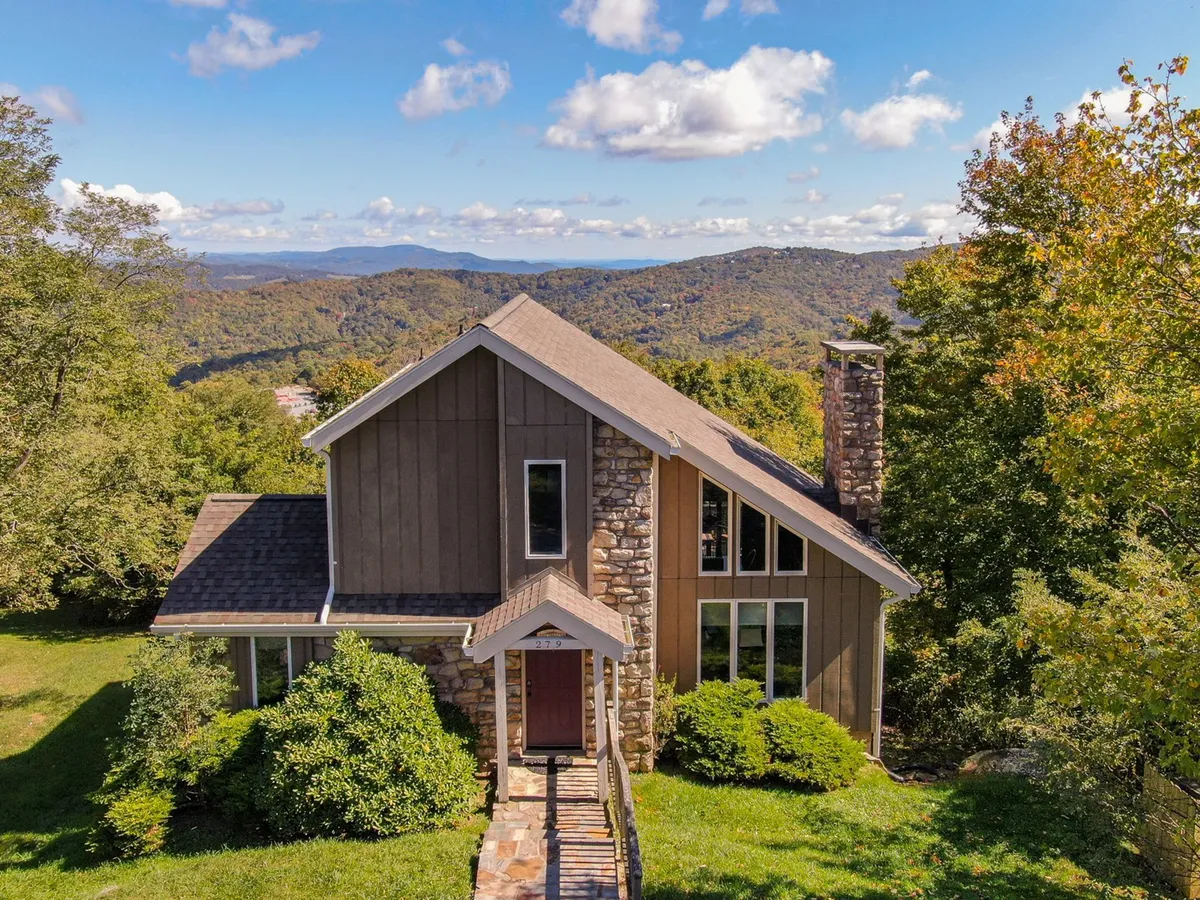 The image features the exterior of a beautiful three-story mountain home, showcasing a striking stone facade, expansive windows that capture natural light, and a welcoming pathway leading through a lush tree line to the entrance. The setting is serene, with the home nestled in nature, suggesting opportunities for enjoying sunsets from the porch and easy access to nearby hiking trails.