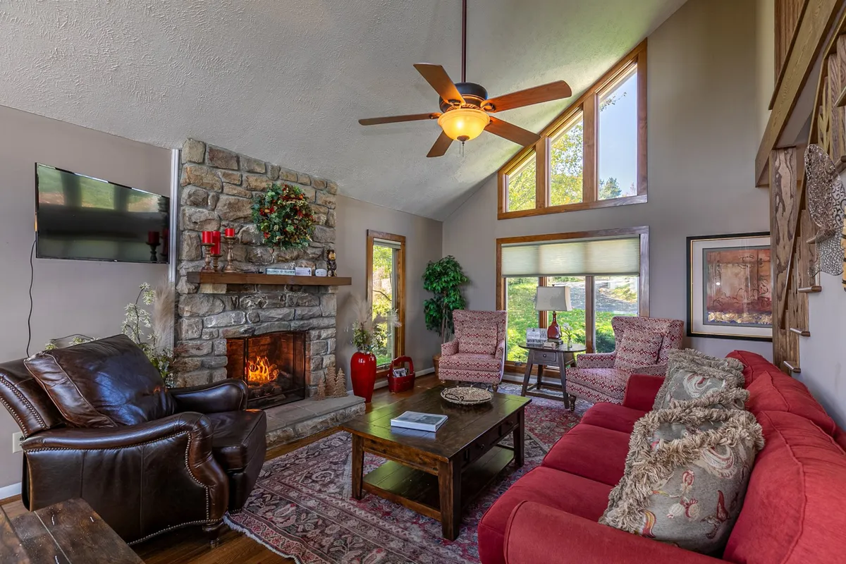 A warm, welcoming living space centered on a stone fireplace with comfortable seating and large windows that fill the room with light. The wooden coffee table suggests a spot for games or snacks, and the windows frame the outdoors, inviting guests to appreciate the scenic setting.