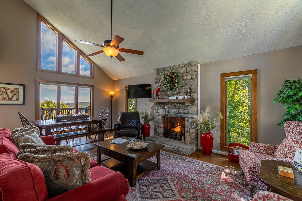 This image showcases the main living room featuring a striking stone fireplace, a red sofa, and a rustic wooden dining table. Large windows allow natural light to stream in, creating a bright and welcoming atmosphere. The space is arranged for gatherings, with cozy seating inviting relaxation and conversation.