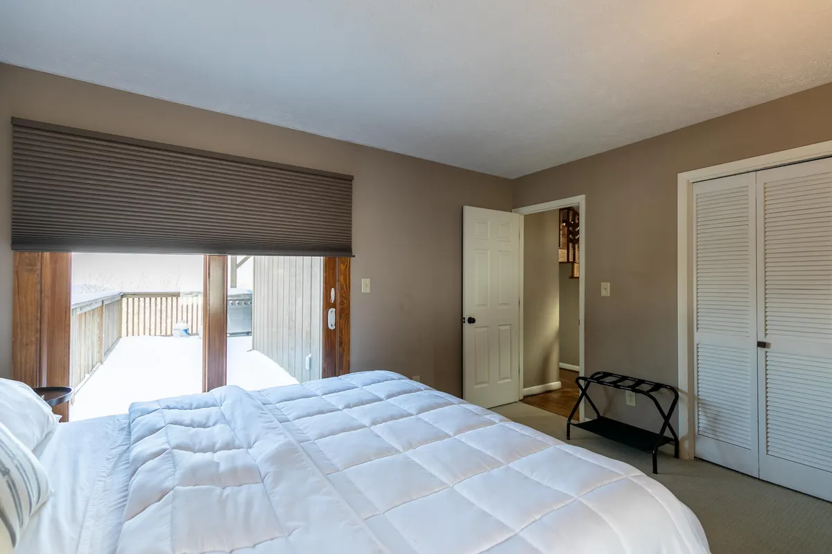 A serene bedroom with a crisp white comforter on the bed faces a large window, which offers a clear view of the outdoors. The gentle sunlight and tranquil atmosphere evoke a sense of peaceful escape and restful mornings amid nature.