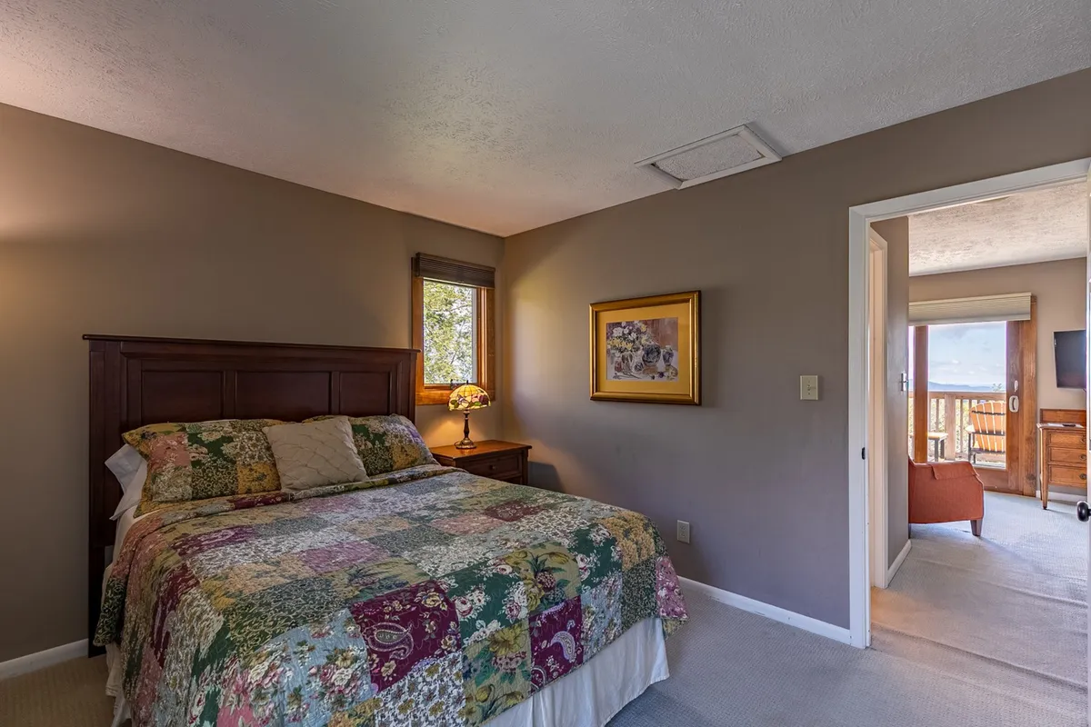 A homey bedroom with a bed topped with a patchwork quilt, a lamp on a bedside table, framed artwork on the wall, and a window letting in gentle natural light. The overall atmosphere is inviting, with a sense of comfort and a personal touch.