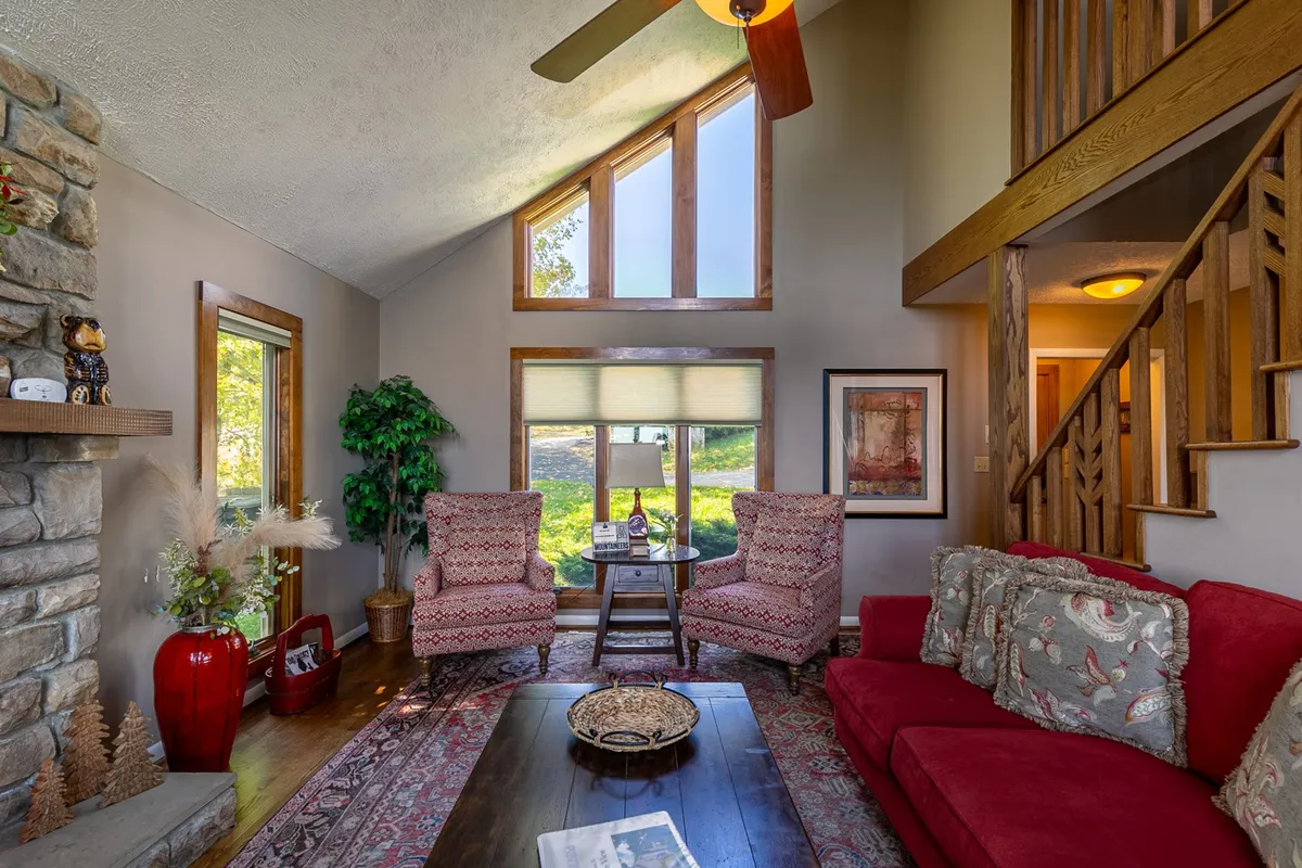 This image captures a spacious interior living area featuring a vibrant red couch, two patterned chairs, and a large window that brings in natural light and offers nature views. A stone fireplace stands as a central gathering spot, blending indoor comfort with a connection to the outdoors.