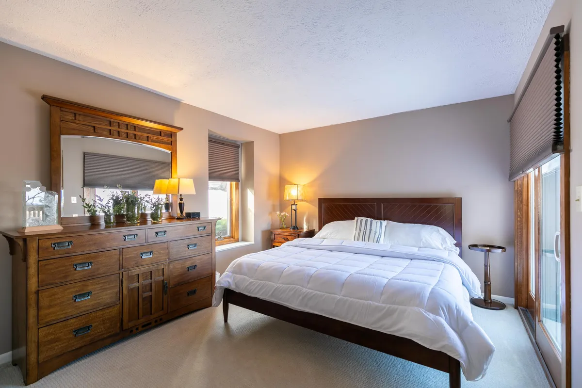 A serene bedroom with a wooden dresser and mirror, a comfortable bed dressed in white bedding, a table lamp for evening reading, and a wooden nightstand. Bright morning light filters in through the window, emphasizing the room's calm and restful mood.
