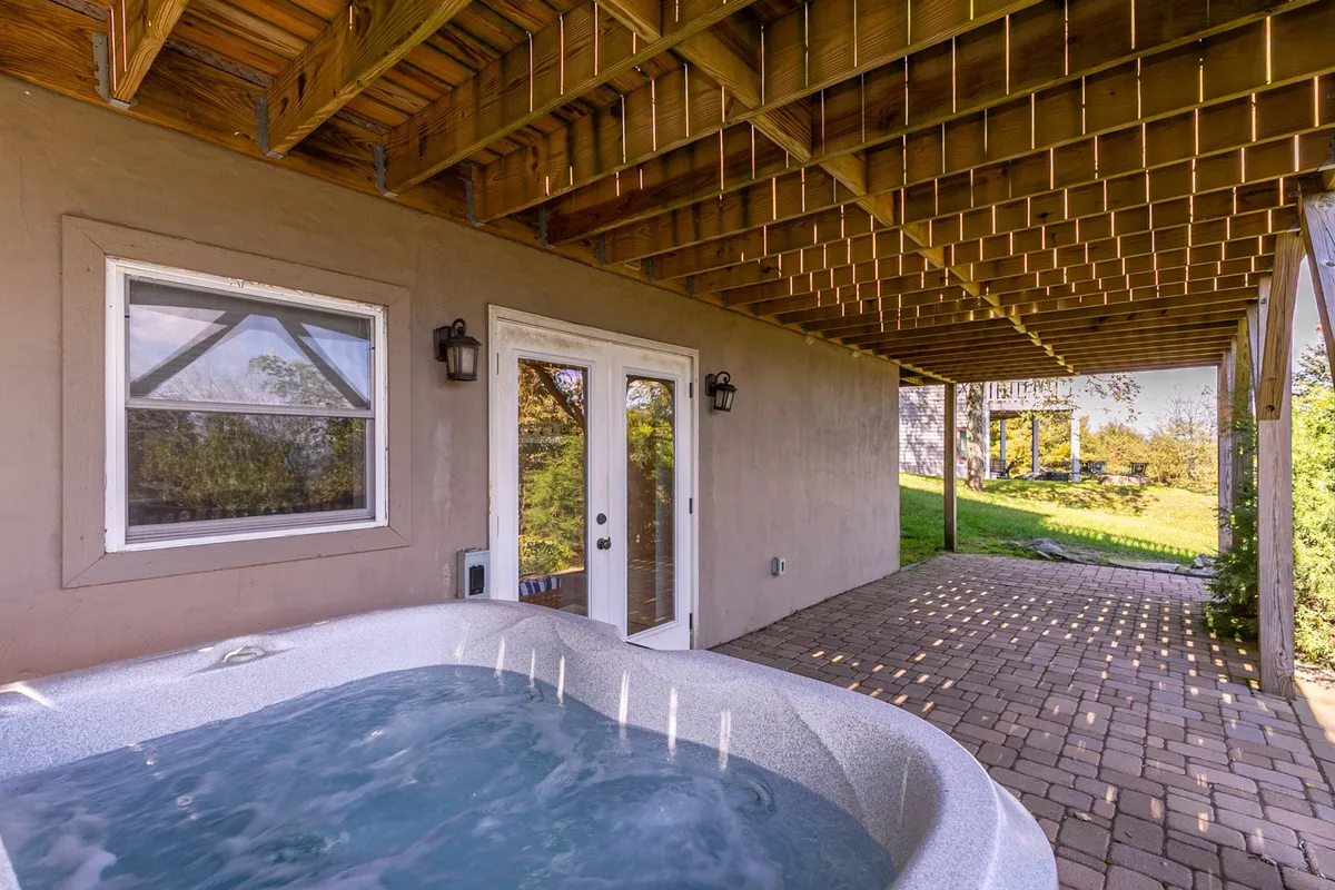 A paved patio with a hot tub positioned near double doors, complemented by outdoor dining space. The area is ideal for relaxation and social gatherings, with the option to move easily between the indoors and outdoors.