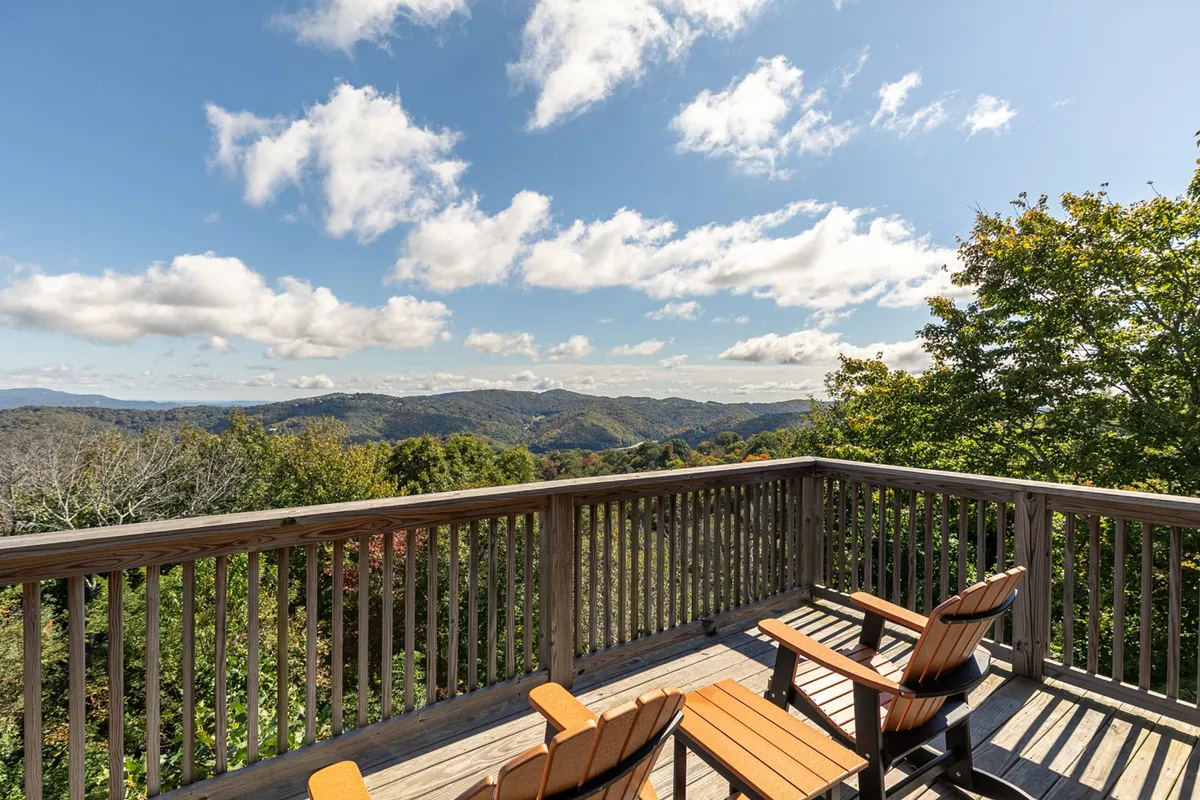 A wooden deck furnished with comfortable outdoor chairs offers a sweeping view of the surrounding mountains. The scene is peaceful, with the last rays of sunlight casting a warm glow, inviting guests to relax and take in the scenery after a day of winter adventure.