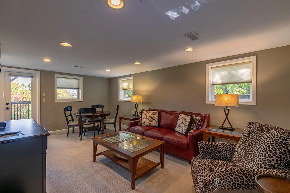 An open-concept interior is displayed, featuring a red leather sofa, a wooden coffee table, a rustic dining table, and a playful leopard print chair. The arrangement encourages socializing and group activities, with space for games, meals, and relaxation.